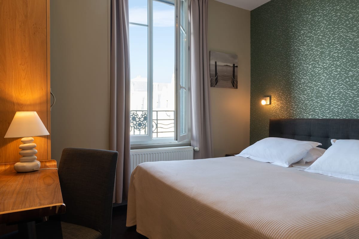 Une chambre d’hôtel comprenant un lit, un bureau avec une lampe, une chaise, une fenêtre avec des rideaux et un mur orné d’un motif texturé.