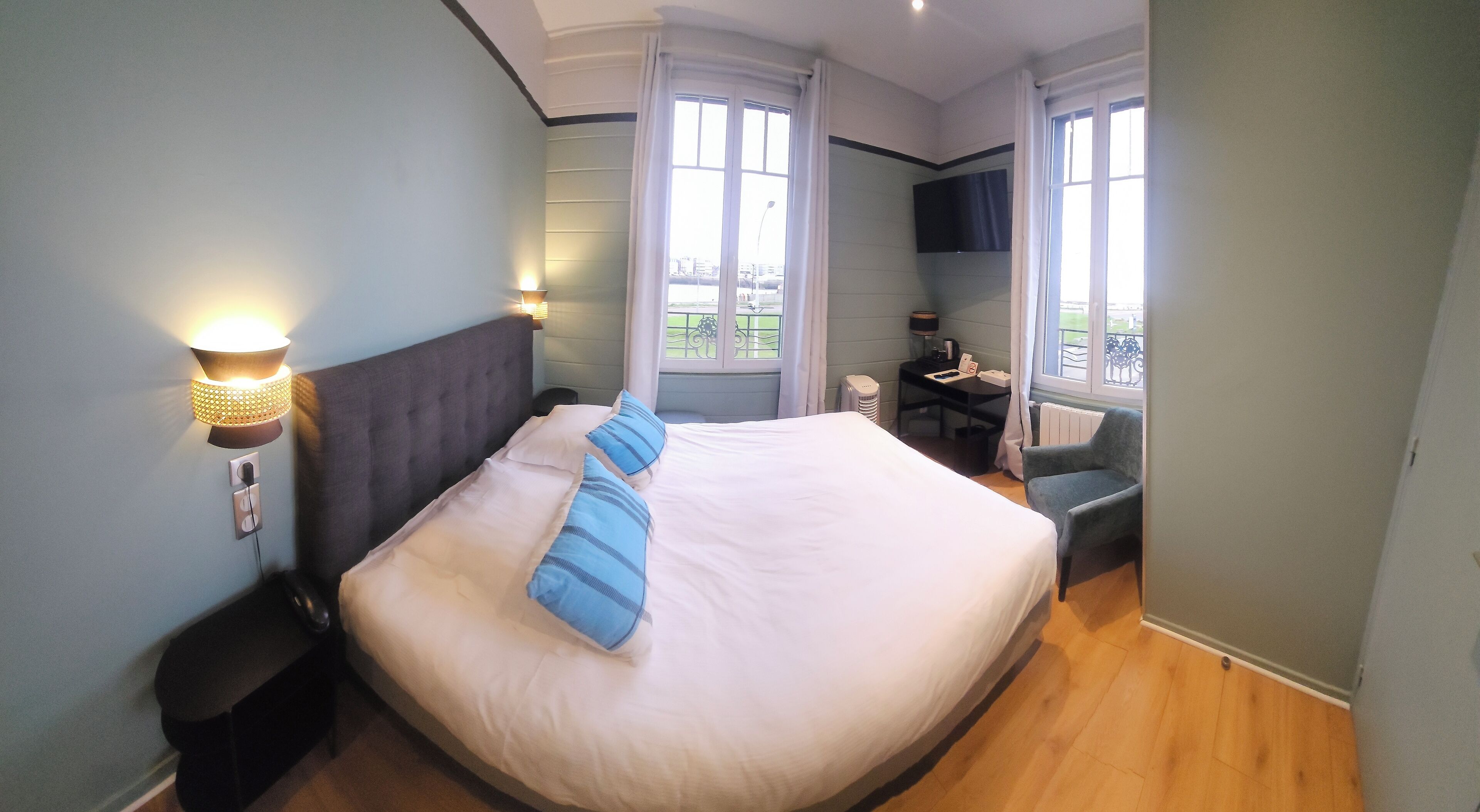 A hotel room with a large bed, a desk with a chair, a TV mounted on the wall, and a window with a view of the outside.