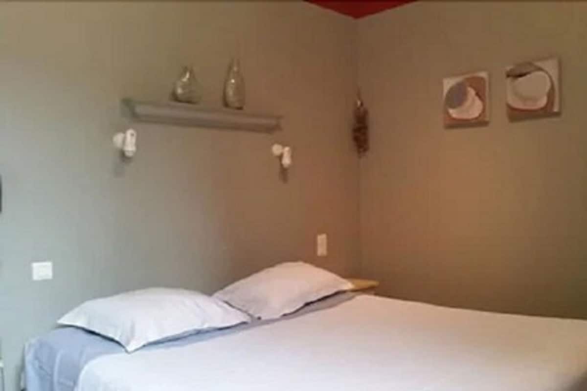 Une chambre à coucher avec un lit, des oreillers, une étagère avec des objets décoratifs et un interrupteur fixé au mur.