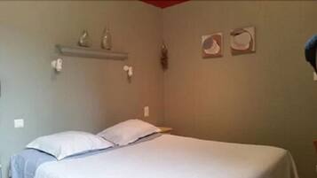 A bedroom with a bed, pillows, a shelf with decorative items, and a wall-mounted light switch.