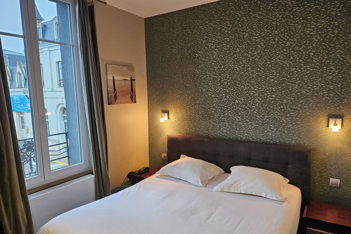 Une chambre d’hôtel avec un grand lit, une table de chevet, un téléphone et une fenêtre avec des rideaux.