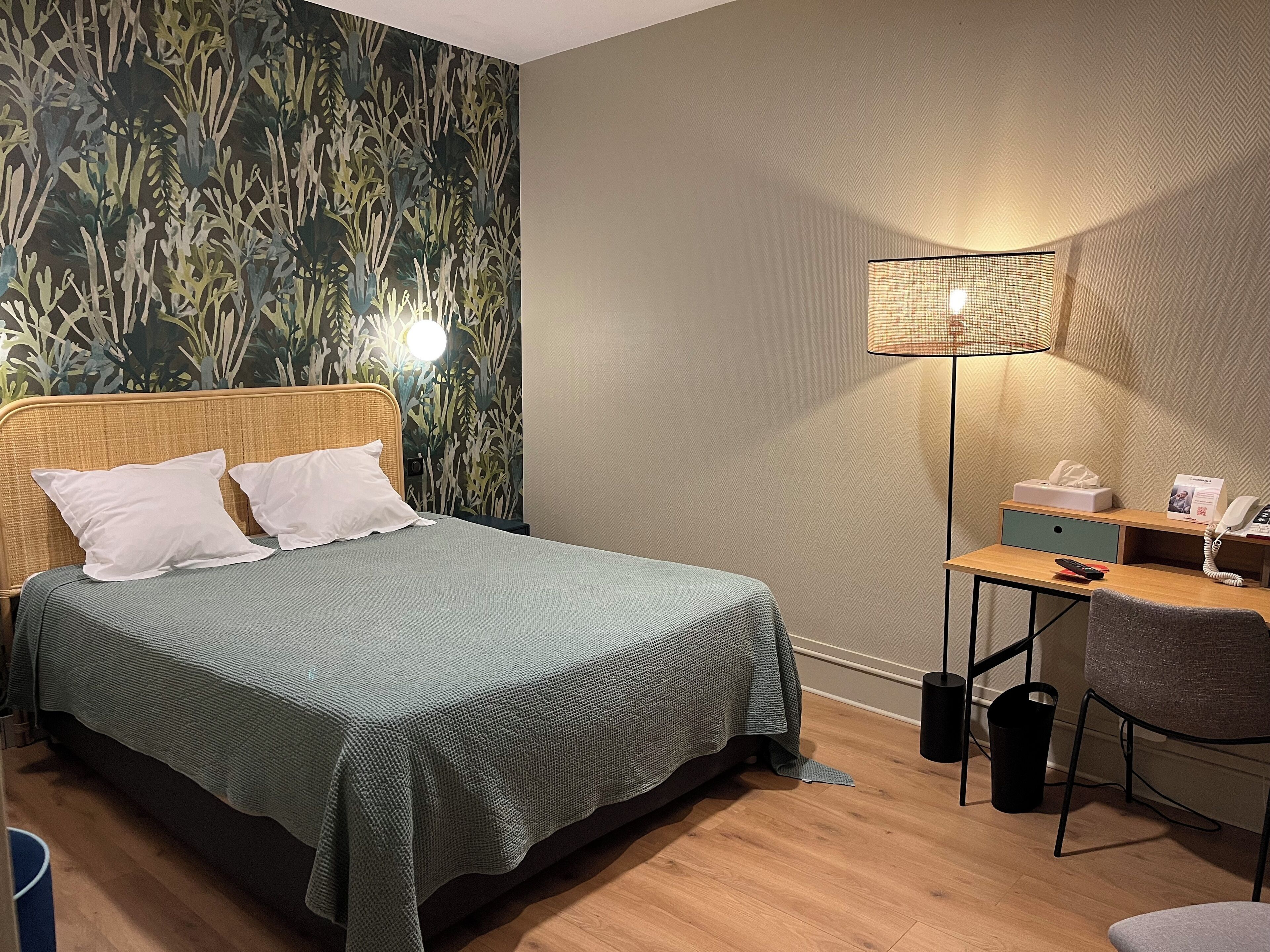 Une chambre d’hôtel comprenant un lit, un bureau, une chaise, une lampe et un mur orné d’un motif végétal.