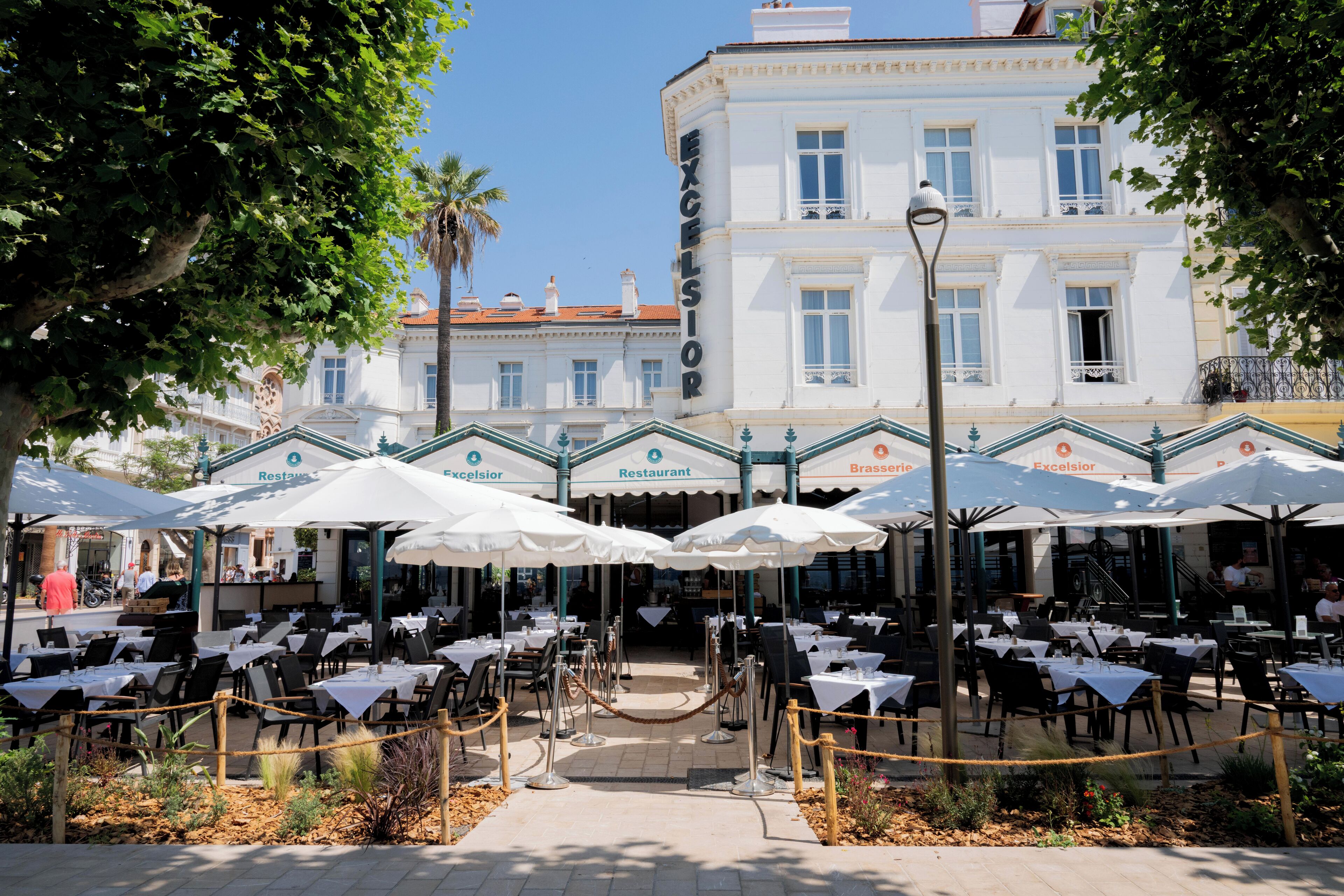 An outdoor dining area with tables and umbrellas, a building with 'Excelsior' signage, and a clear sky.