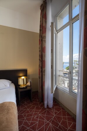 A hotel room with a bed, a nightstand with a lamp, a window with a view of the sea, and a patterned carpet.