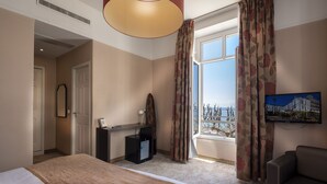 A hotel room with a large bed, a desk with a computer, a chair, a TV mounted on the wall, and a window with curtains. - Excelsior (Saint-Raphael)
