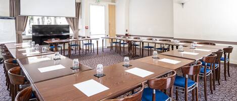 A conference room with long tables, chairs, a projector screen, and a television.