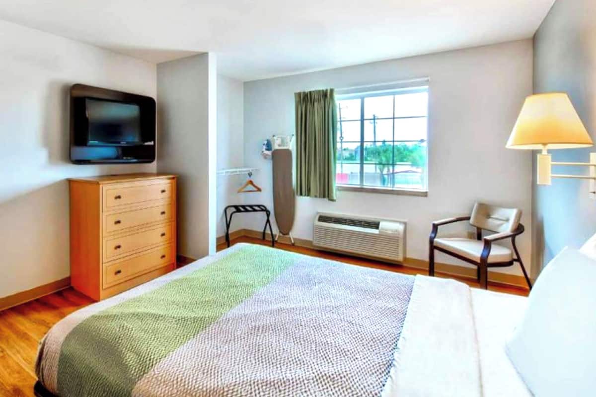 Camera d'albergo con un letto, una cassettiera, una televisione a parete, una finestra con tende, una sedia e una lampada.