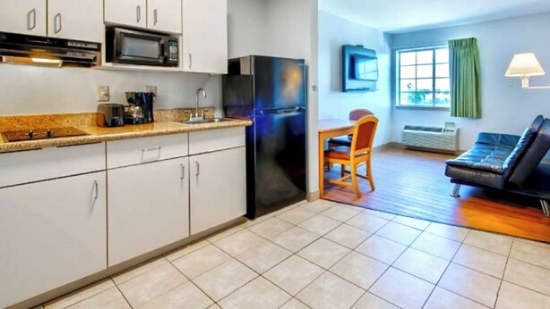 A compact kitchen with white cabinets, a microwave, and a black refrigerator. There's a dining area with a wooden table and chairs, and a black sofa. - Oyo Townhouse Sulphur,LA Extended Stay I-10 (Sulphur)