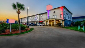A hotel with a central entrance, multiple parking spaces, and a landscaped area with palm trees. - Oyo Townhouse Sulphur,LA Extended Stay I-10 (Sulphur)