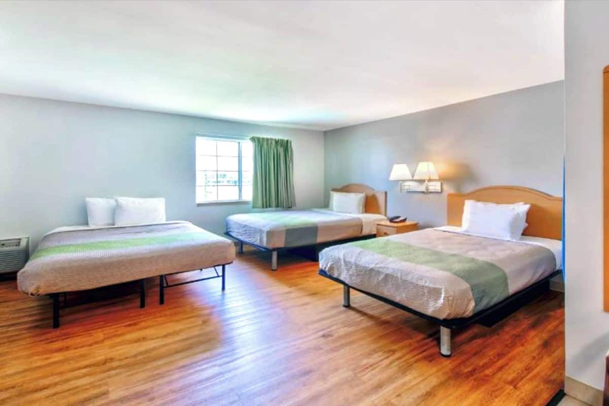 Una camera d'albergo con tre letti, pavimento in legno, una finestra con tende e lampade da comodino.