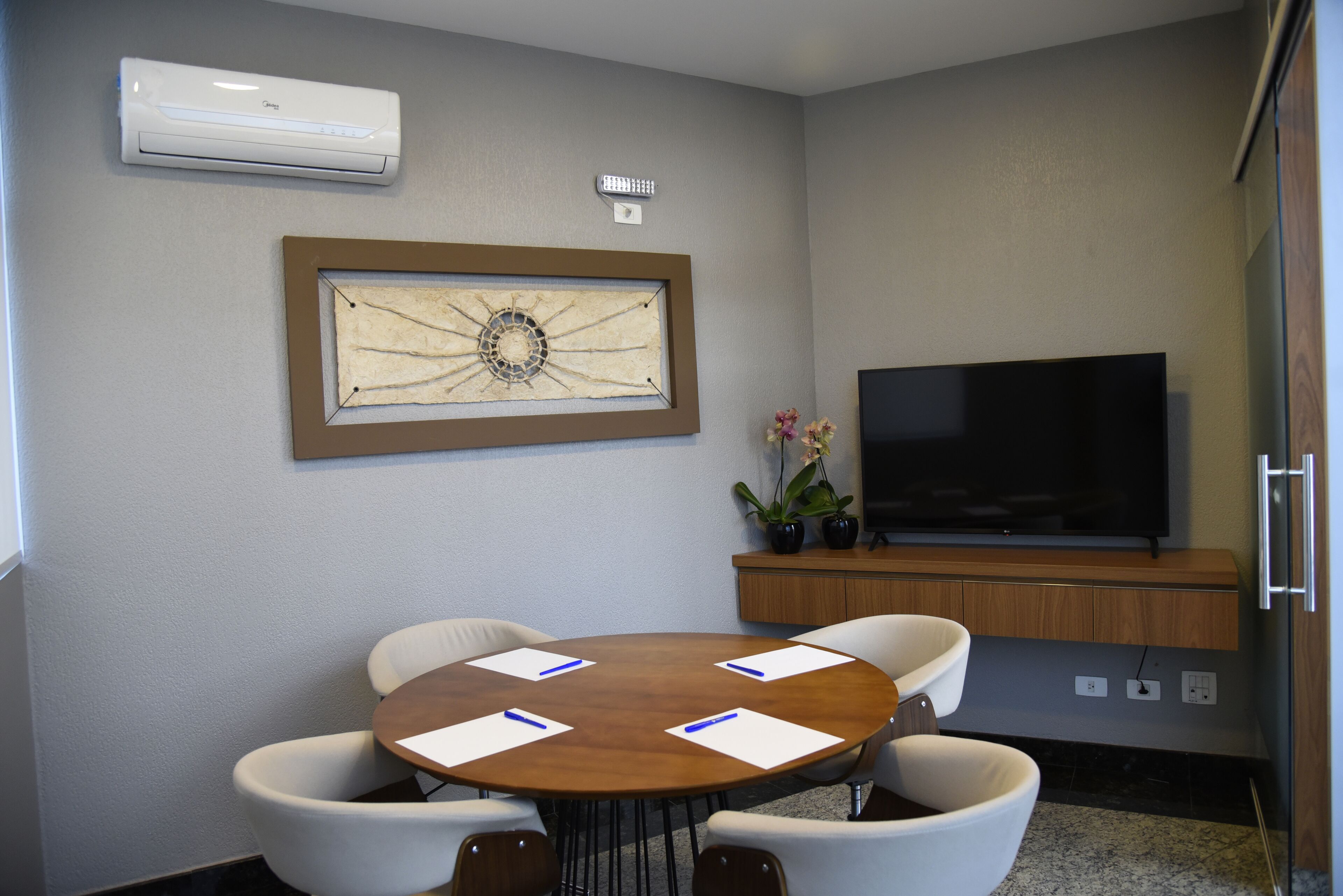 Uma sala de conferências com uma mesa redonda, cadeiras brancas, uma obra de arte emoldurada, uma televisão e um aparelho de ar condicionado.