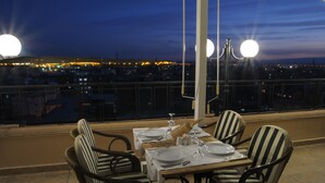 A dining table set for four with striped chairs, a city view at dusk, and a railing in the foreground. - SV Business Hotel Diyarbakır (Diyarbakir)