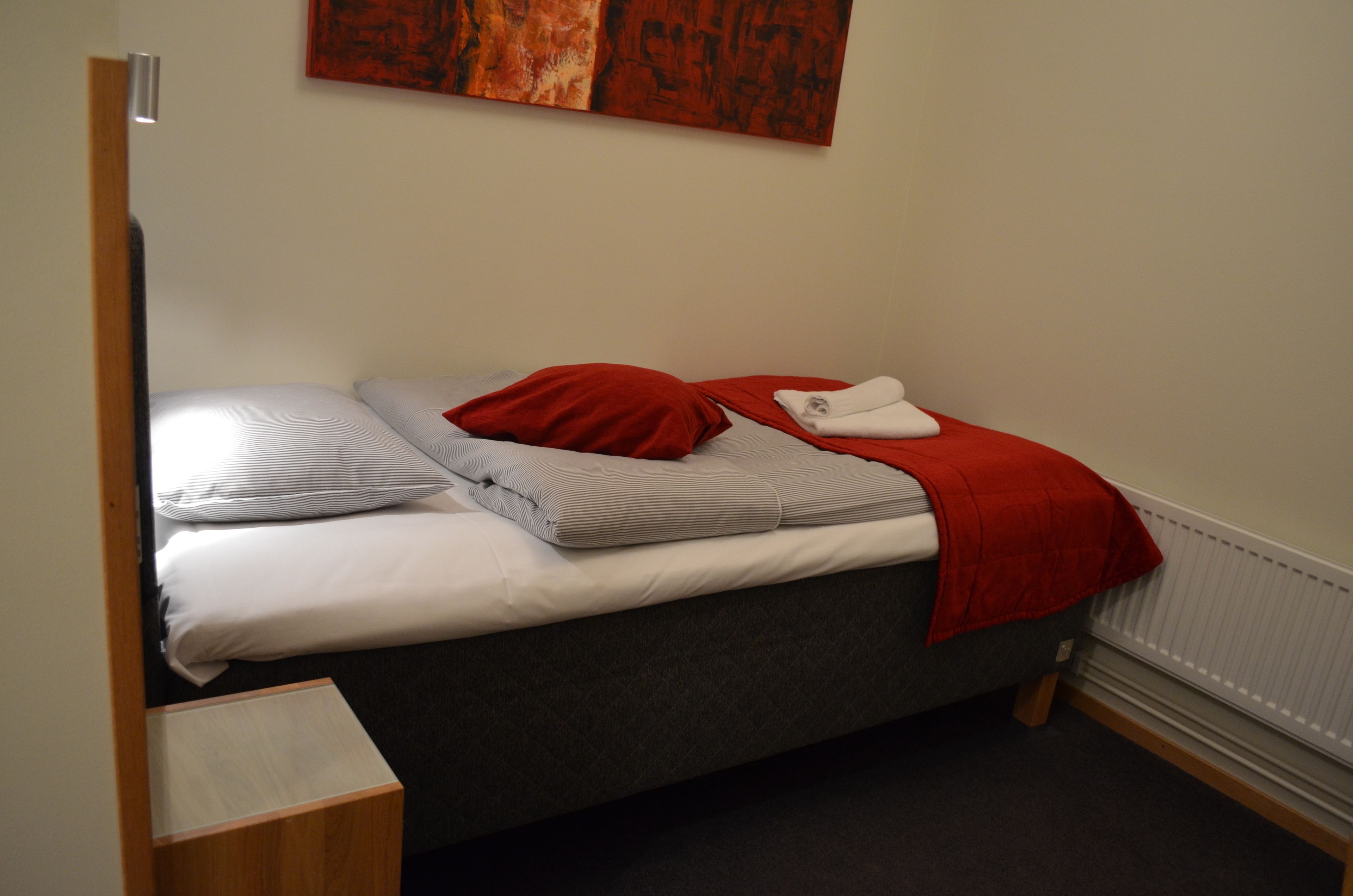 Una cama individual con colchón gris, almohadas blancas y grises, una colcha roja y una toalla blanca.