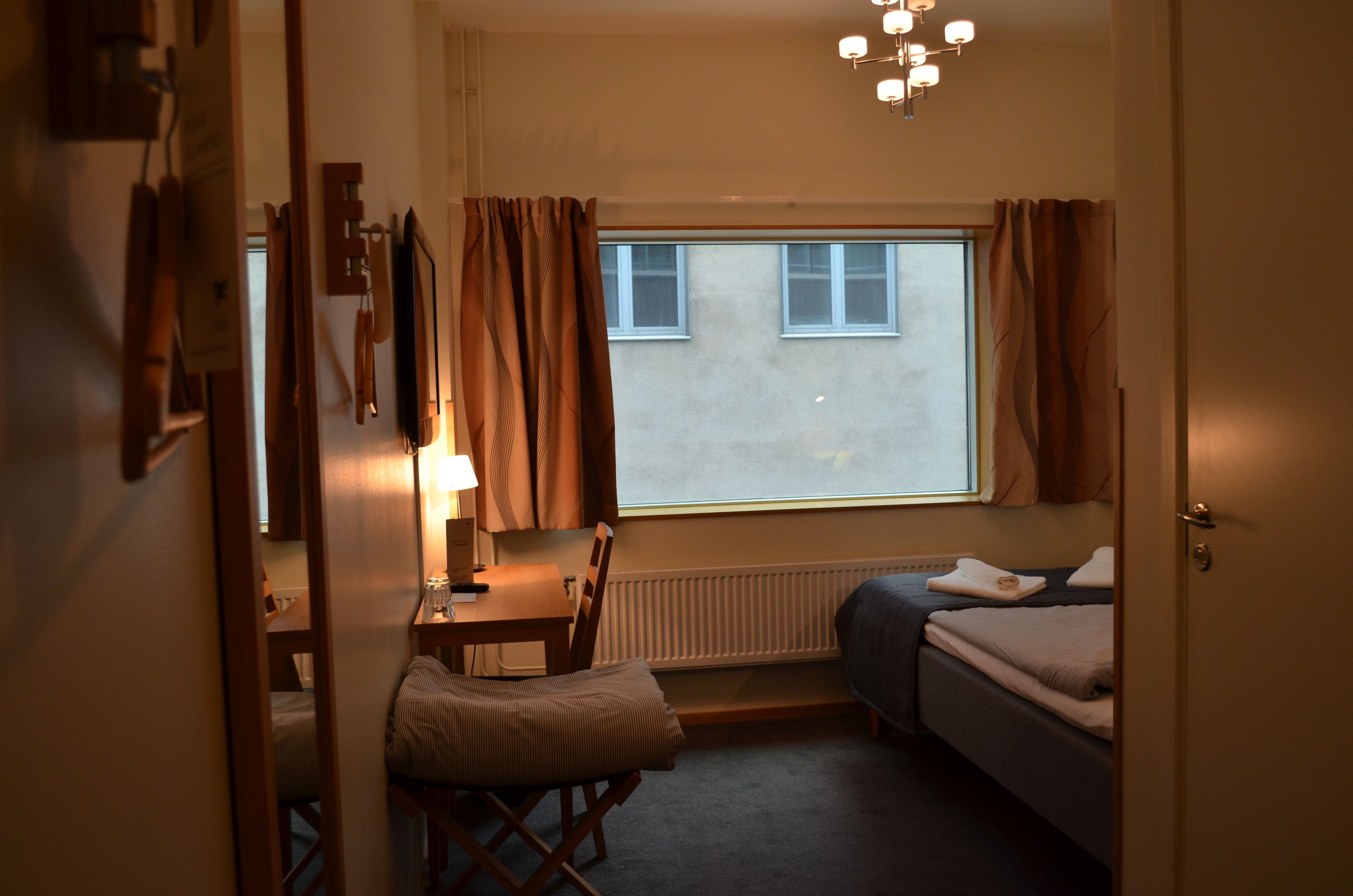 Et hyggeligt, lille værelse med seng, skrivebord og stol. Et vindue med gardiner, en radiator og en loftlampe.
