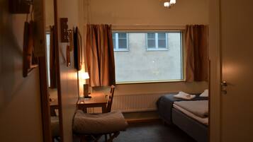 Et hyggeligt, lille værelse med seng, skrivebord og stol. Et vindue med gardiner, en radiator og en loftlampe.