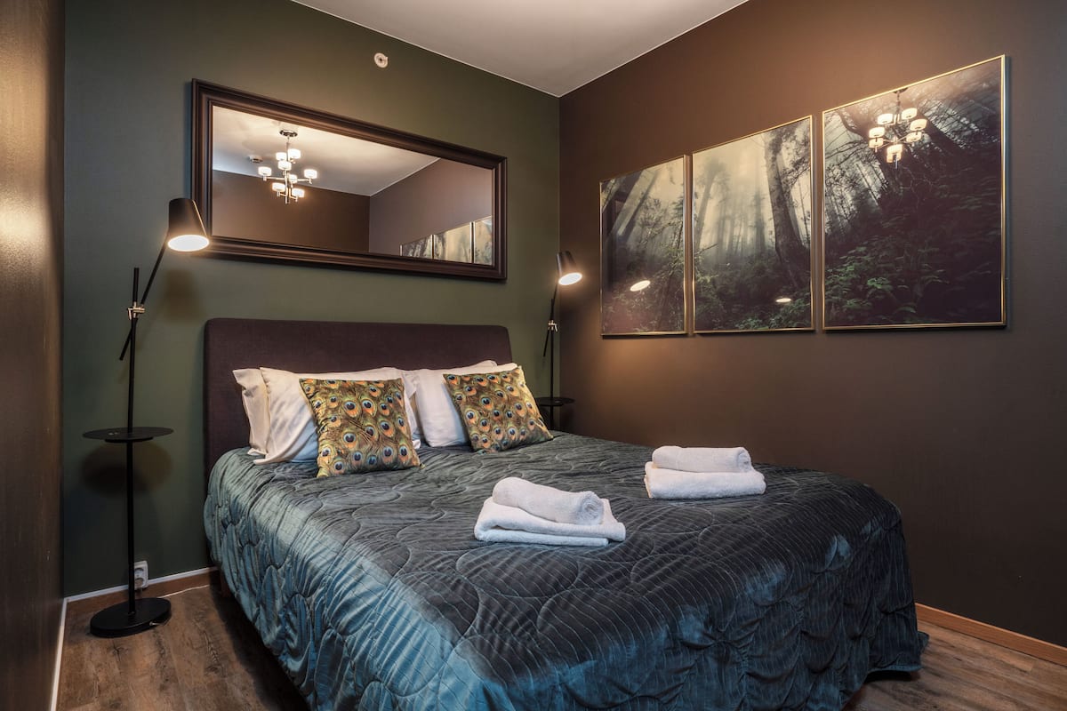 A bedroom with a large bed, two bedside lamps, a mirror, and a forest-themed wall art.