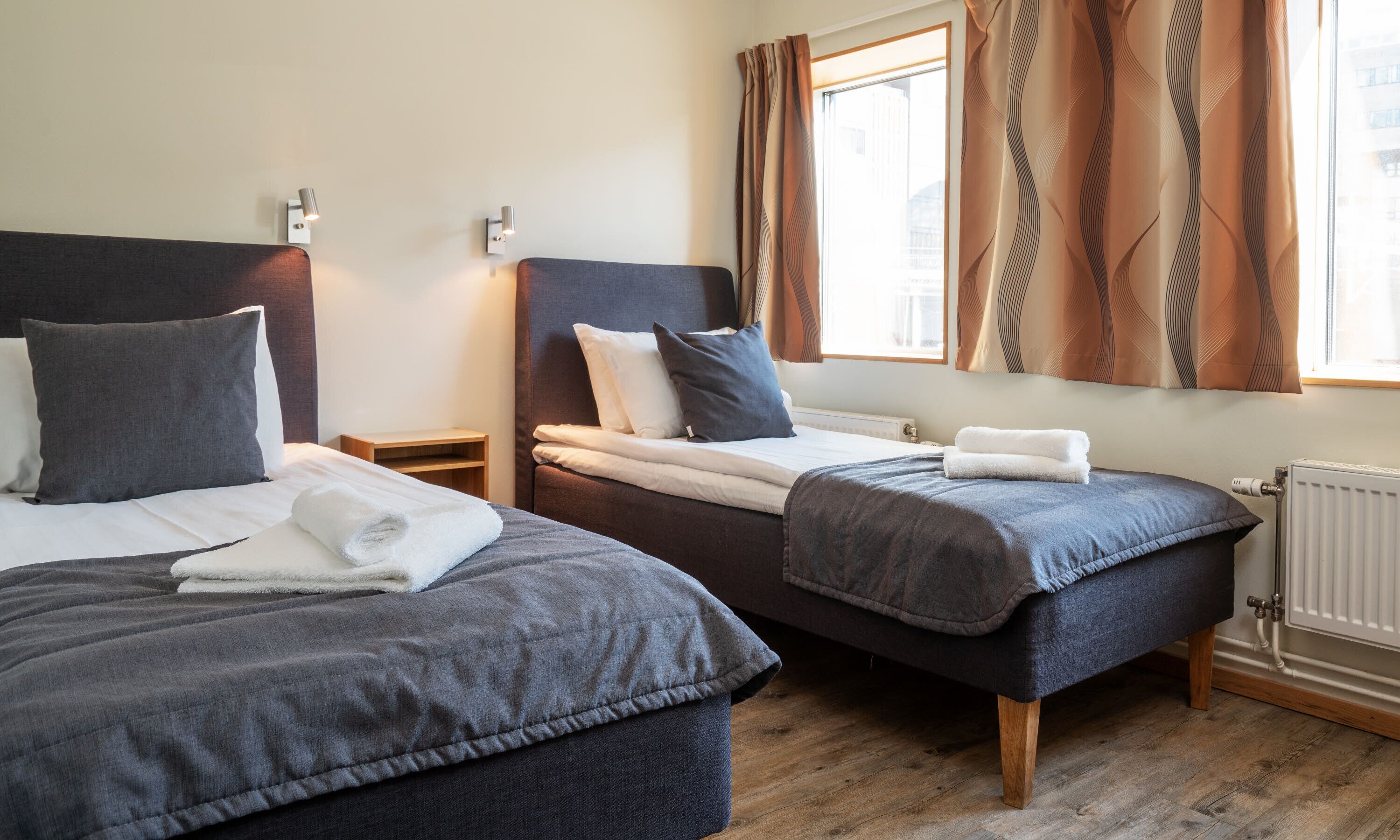 Habitación de hotel con dos camas, cada una con almohada y toalla doblada. Entre las camas hay una mesita de noche de madera, y una ventana con cortinas.