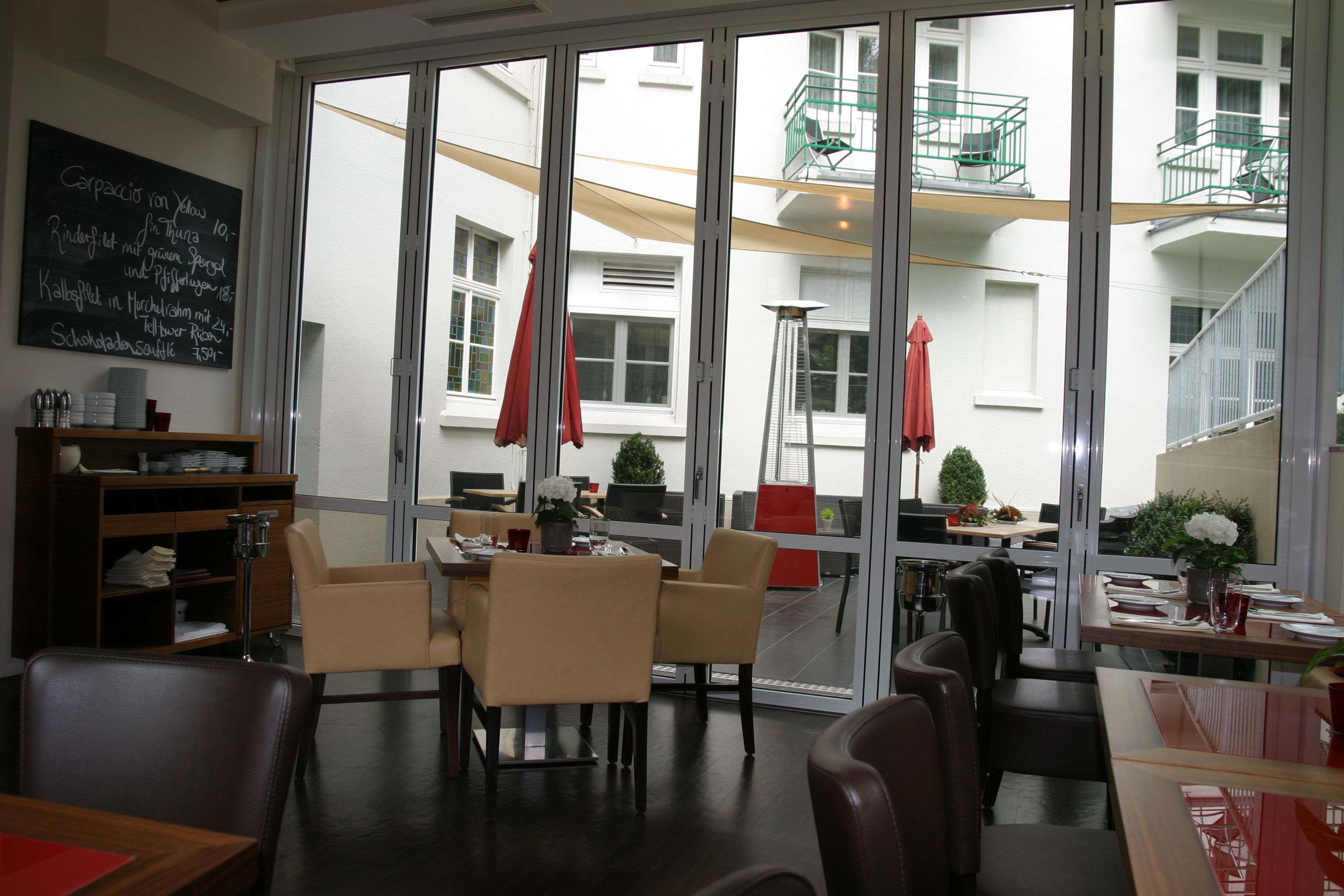 En restaurang med stora fönster, sittplatser inomhus och utsikt över en byggnad med balkonger.