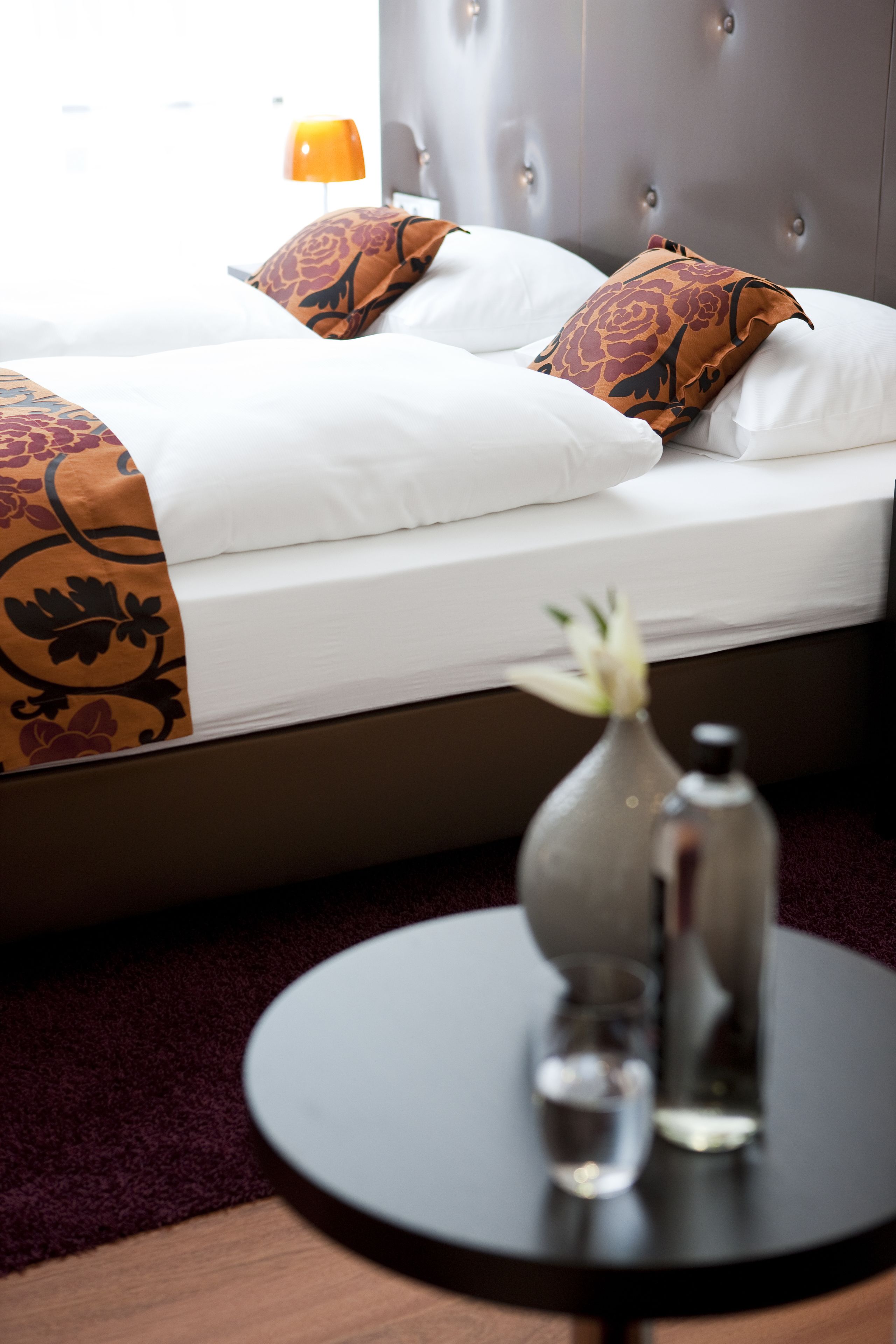 A neatly made bed with white linens and decorative pillows, a vase with a flower, and a small round table with a glass and a bottle.