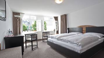 A hotel room with a large bed, a desk with a chair, a window with curtains, and a view of buildings outside.