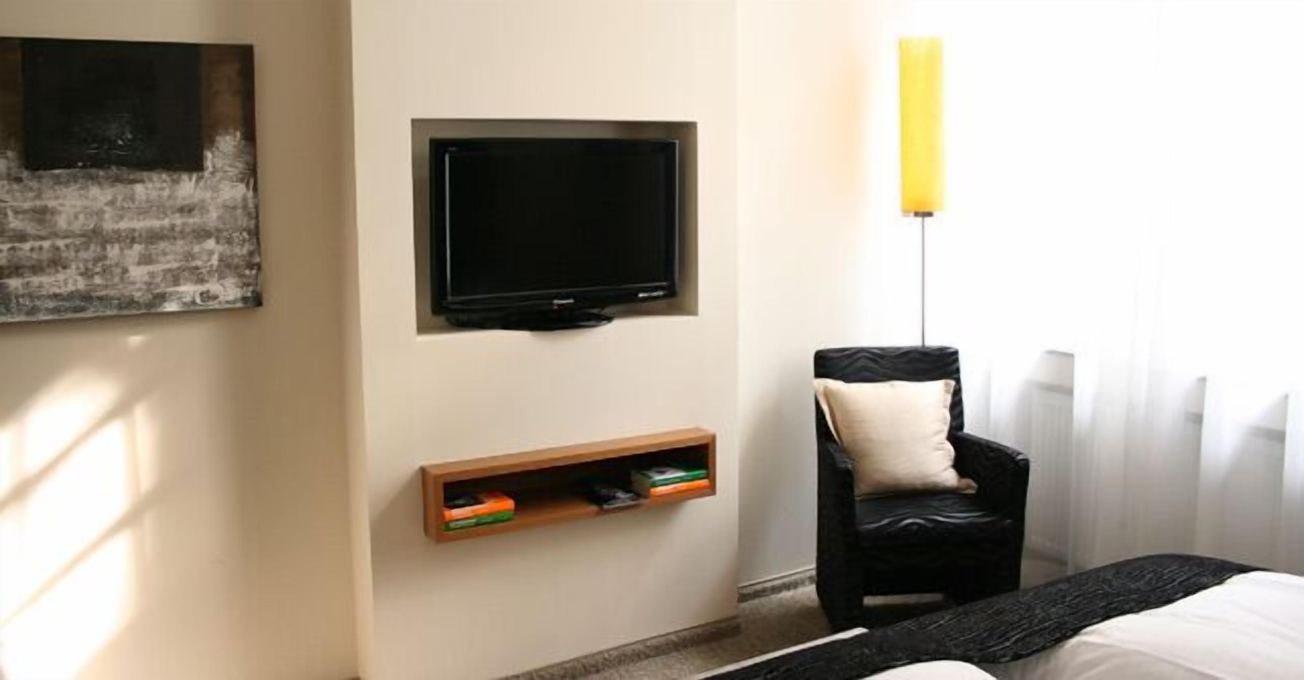 Une chambre d’hôtel dotée d’un téléviseur à écran plat fixé au mur, d’une étagère en bois en dessous, d’un fauteuil en cuir noir avec un coussin et d’un lit recouvert d’un plaid noir.