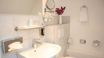 Un baño moderno con lavamanos, espejo y retrete blancos. Hay artículos de aseo en el lavamanos y una toalla colgada en la pared.