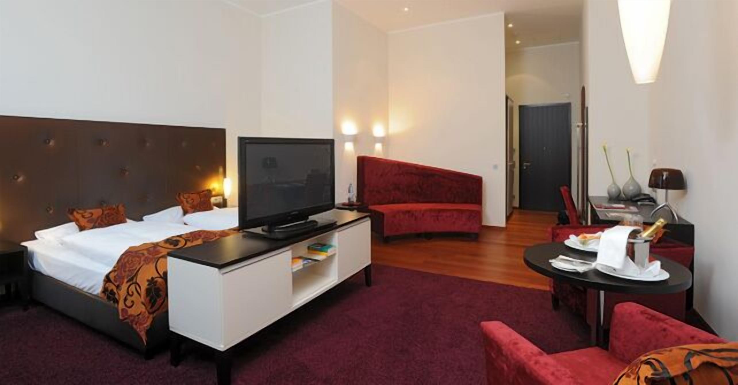 Ein Hotelzimmer mit einem Bett, einem Fernseher auf einer weißen Kommode, einem roten Sofa, einem kleinen Esstisch und einem Spiegel.