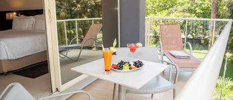 Une chambre d’hôtel avec un balcon, un lit, une table avec des fruits et des boissons, et une chaise.