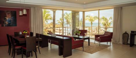 Amplia sala de estar con comedor, sofá y un amplio ventanal con vista a la playa y palmeras.