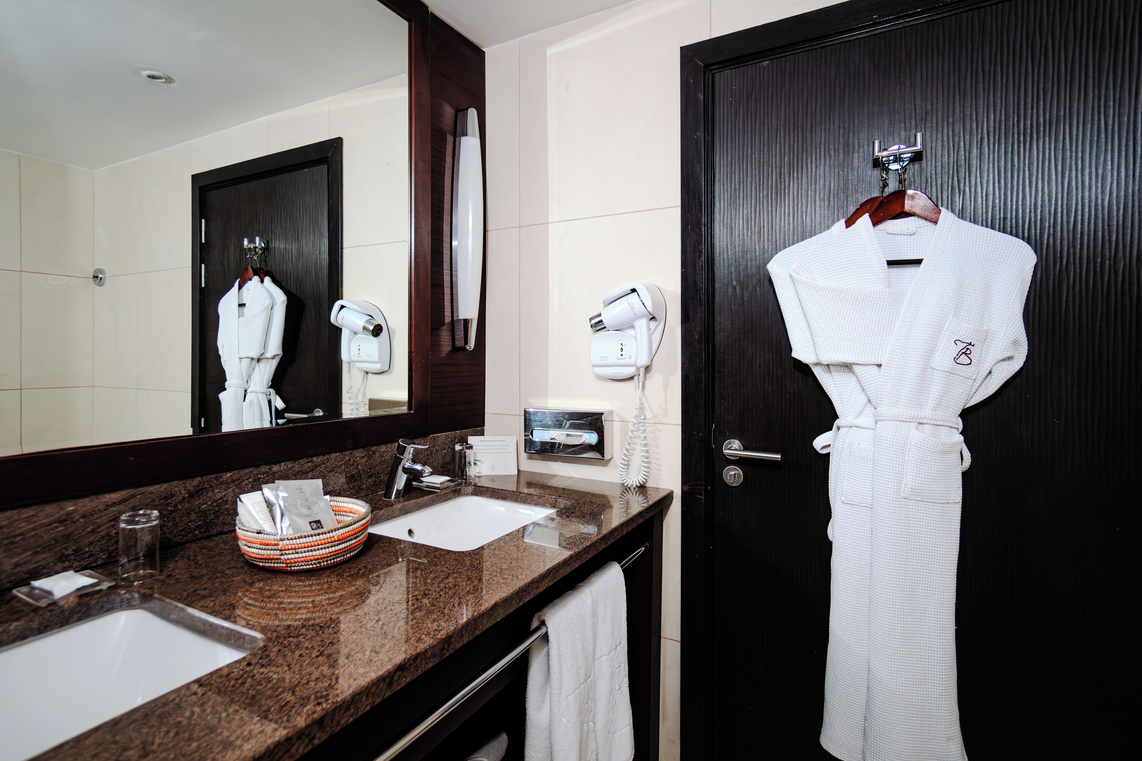 A hotel bathroom with a dark wooden door, a large mirror, a granite countertop, a white sink, a towel rack, and a robe hanging on the wall.