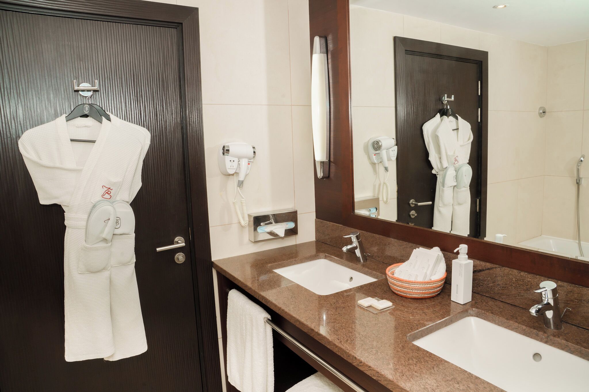 A hotel bathroom with two sinks, a large mirror, a robe hanging on the door, and a towel rack.