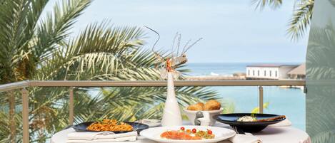 A table set with plates of food, a vase with dried plants, and a view of the ocean and palm trees.