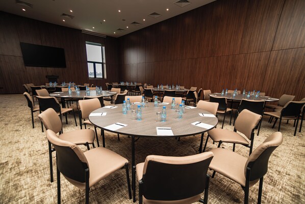 A conference room with multiple tables and chairs, equipped with water bottles and papers, in a modern setting with a large screen and a window.