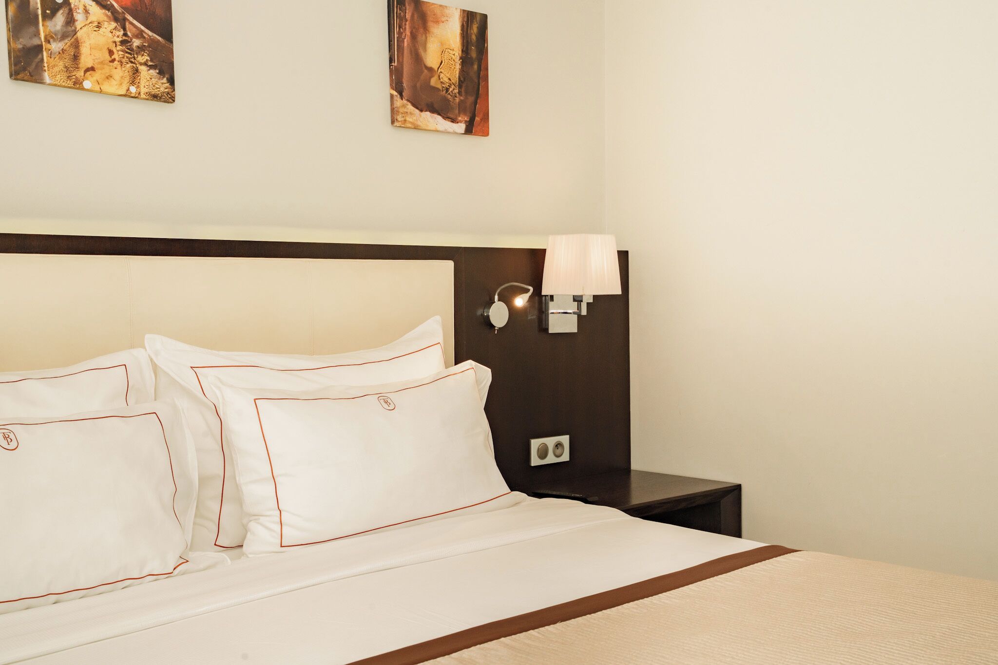 A neatly made bed with white linens and red piping, a dark wooden headboard, and a wall-mounted lamp.
