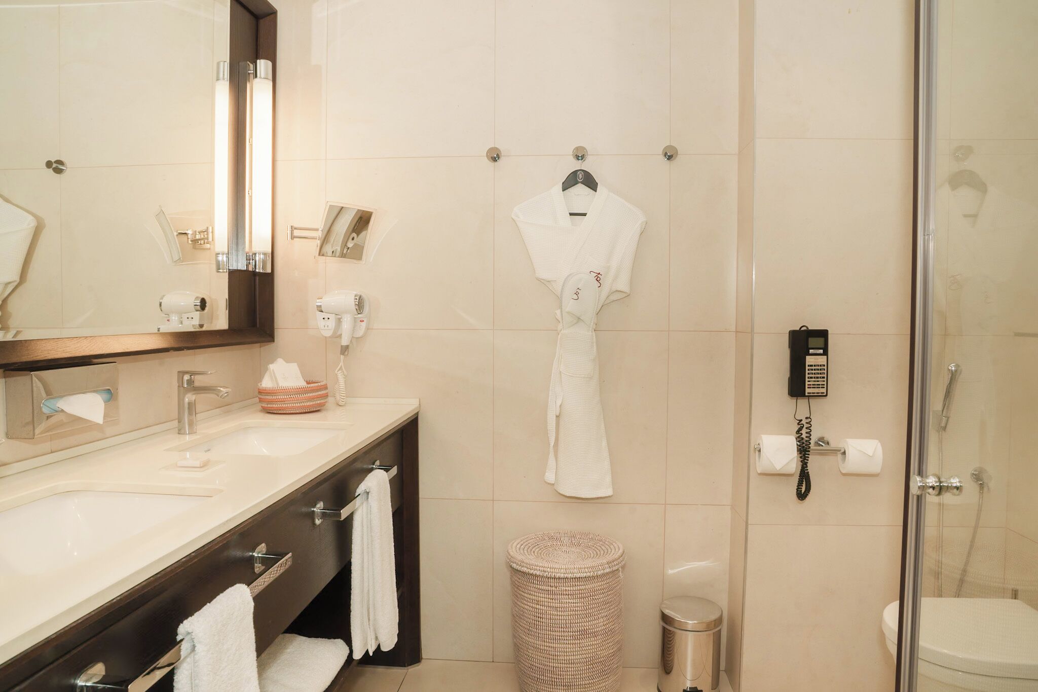A modern bathroom with a large mirror, a sink, a robe hanging on a hook, a shower area, and a waste bin.