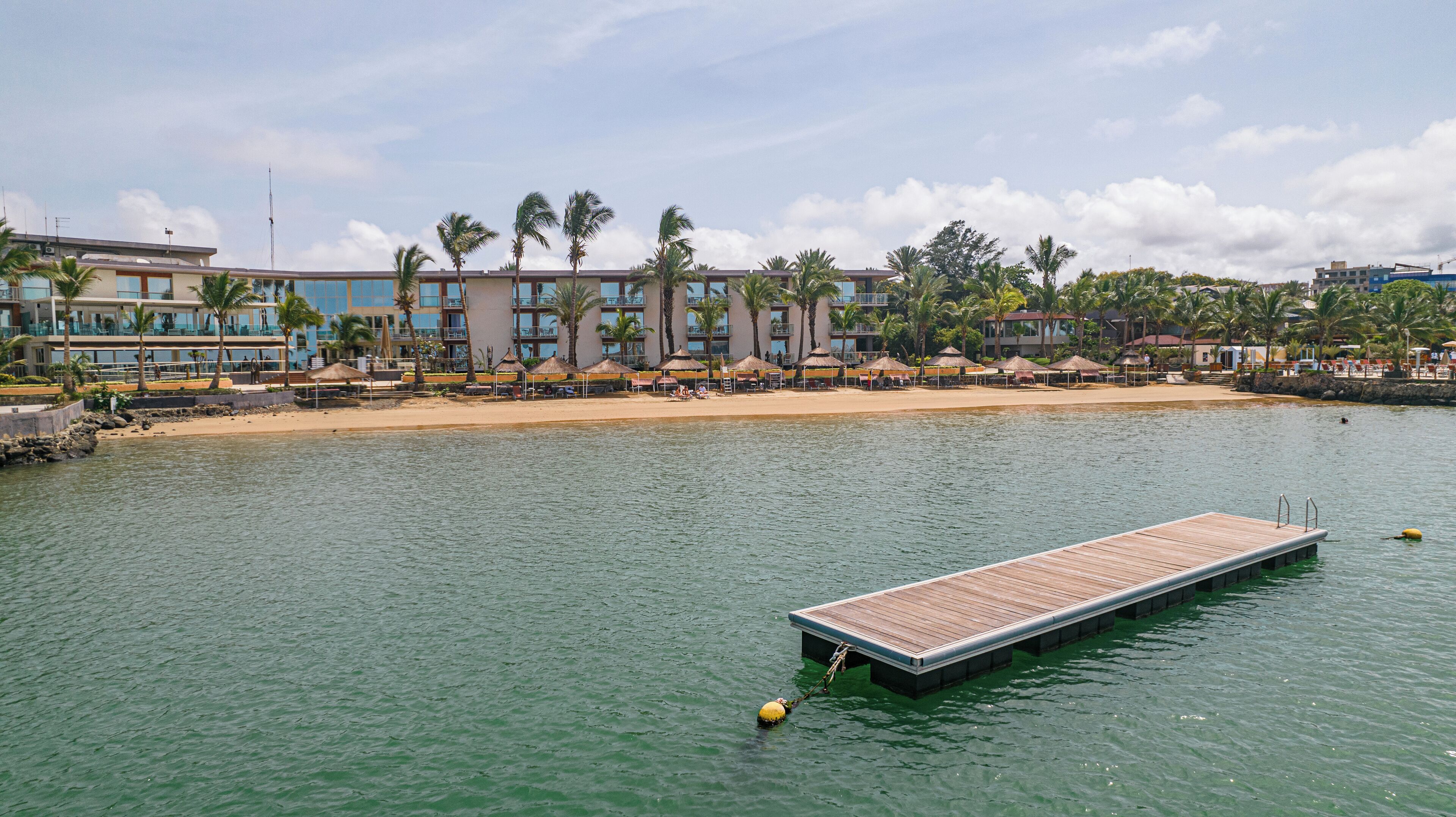 A waterfront hotel with a wooden dock, palm trees, and a sandy beach.