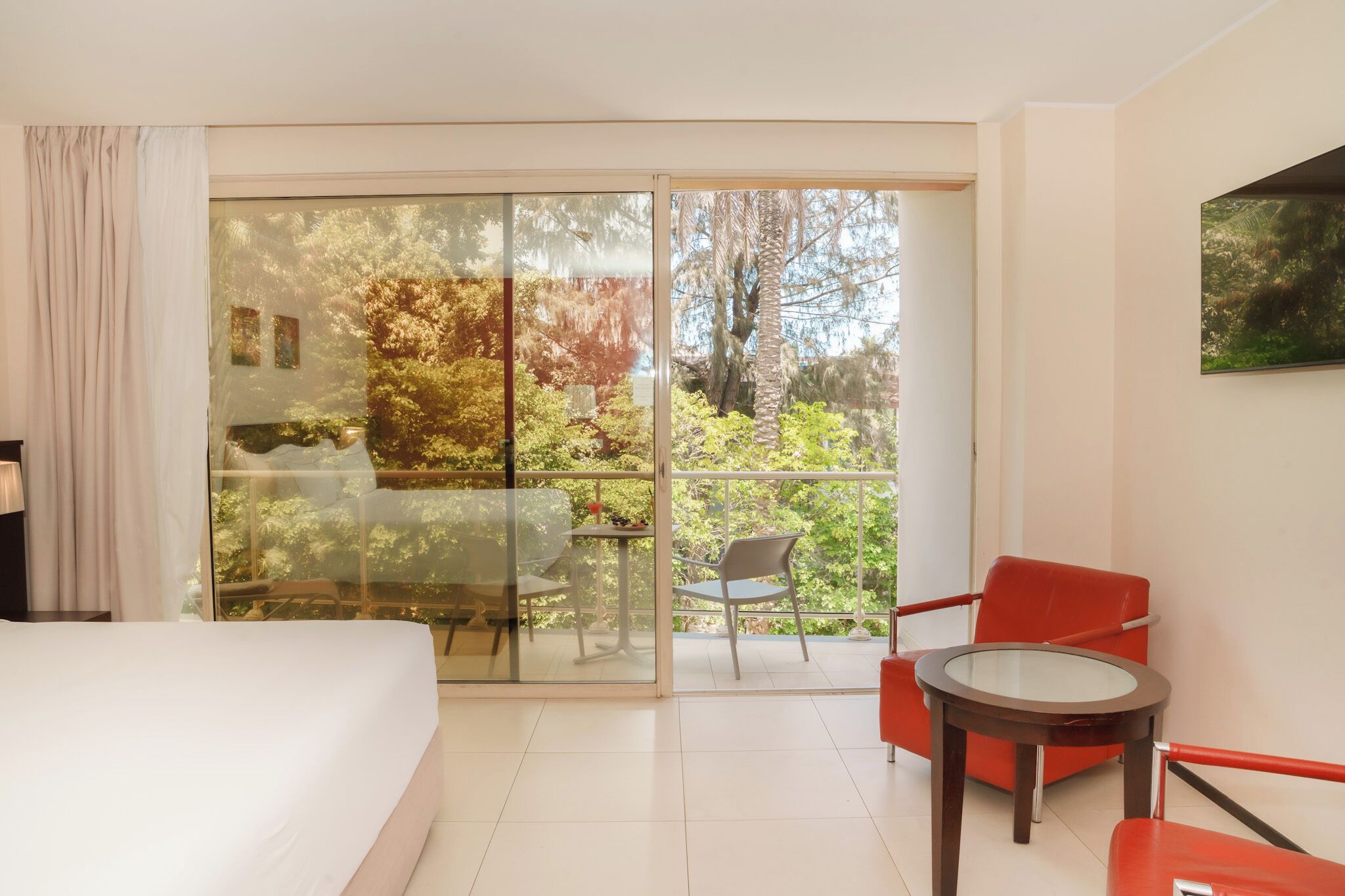 Habitación de hotel con un amplio ventanal corredero que da a un balcón con vistas a árboles y una mesa con sillas.