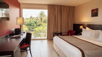 A hotel room with a bed, desk, chair, and a balcony with a view of greenery.