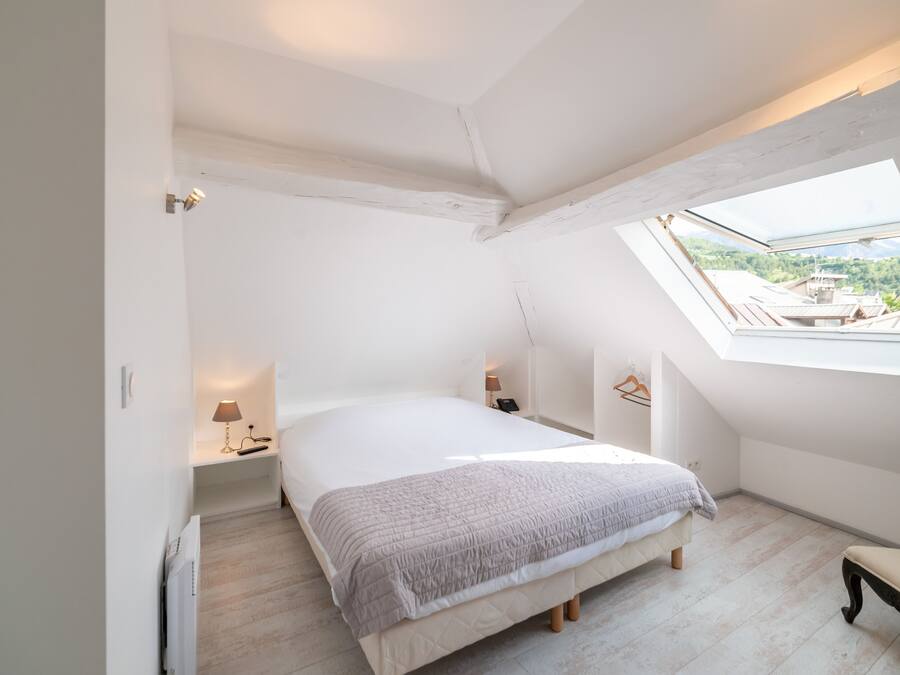 Quarto com claraboia, cama, mesas de cabeceira e um banco.