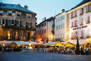 Una plaza animada con mesas al aire libre, una fuente y edificios con letreros de un hotel y una cafetería.