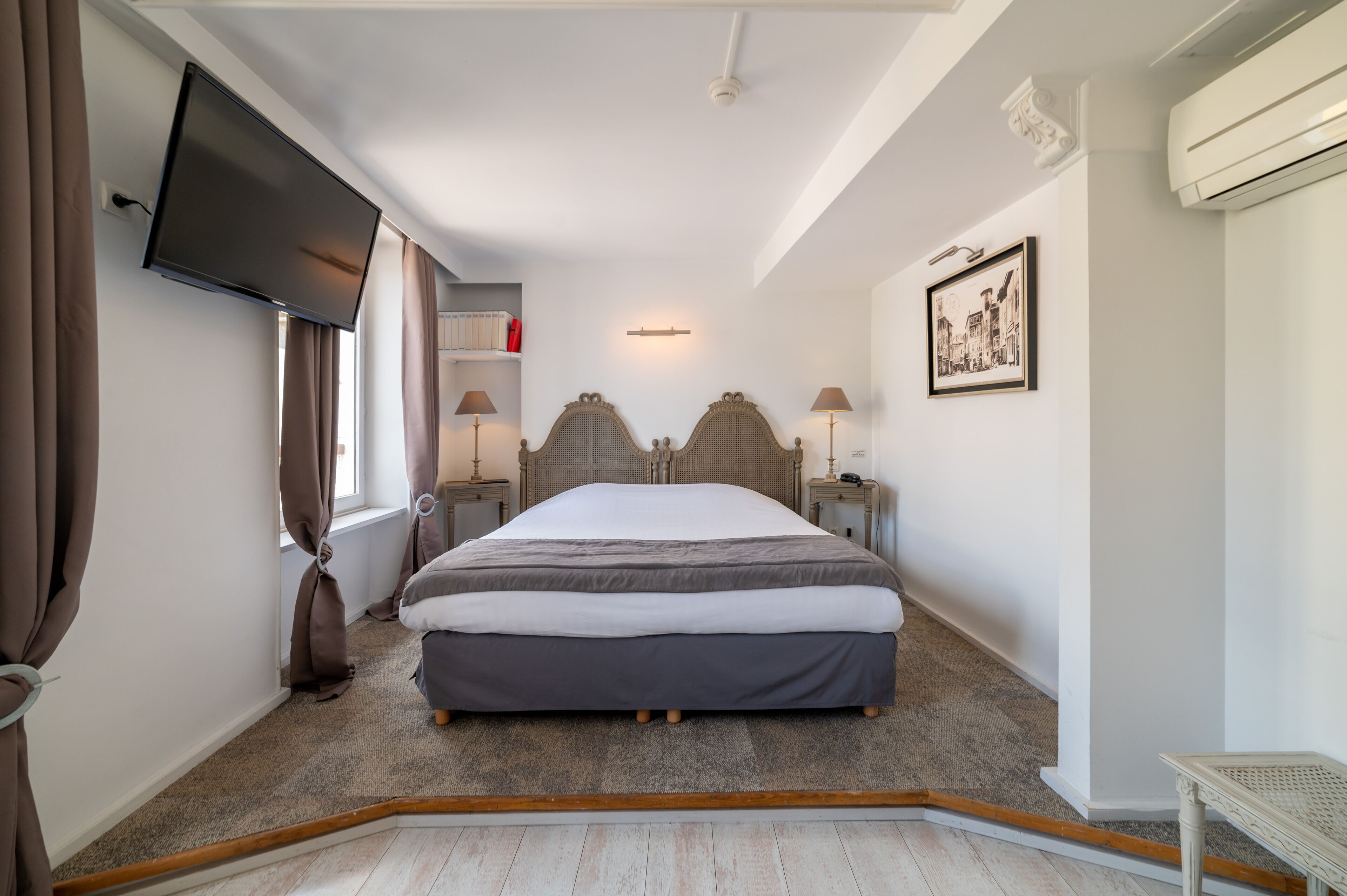 A bedroom with a bed, a flat-screen TV mounted on the wall, a window with curtains, bedside lamps, a framed picture on the wall, and an air conditioning unit.
