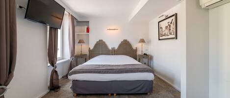 Un dormitorio con una cama, un televisor de pantalla plana montado en la pared, una ventana con cortinas, lámparas de noche, un cuadro enmarcado en la pared y un equipo de aire acondicionado.