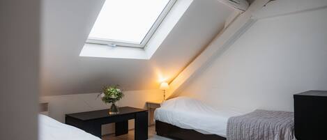 Une petite chambre bien éclairée, dotée d’une lucarne, d’une table de chevet avec une lampe et d’un lit recouvert d’une couverture.
