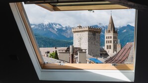 Vista a través d'un òcul de bou d'una torre històrica amb agulla, envoltada de muntanyes i una vall boscosa.