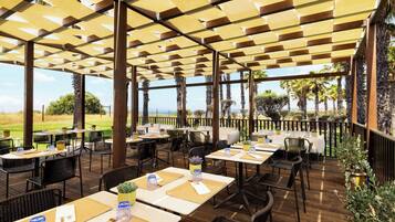 An outdoor dining area with tables and chairs, covered by a wooden pergola, overlooking a scenic view.