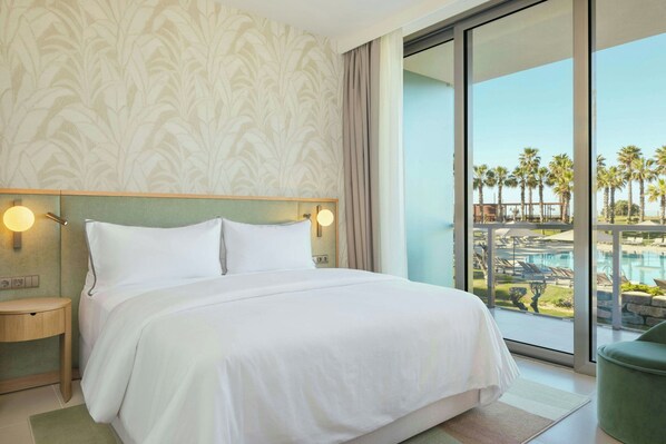 A hotel room with a large bed, a nightstand, a green chair, and a view of a pool and palm trees. - The Westin Salgados Beach Resort, Algarve (Albufeira)
