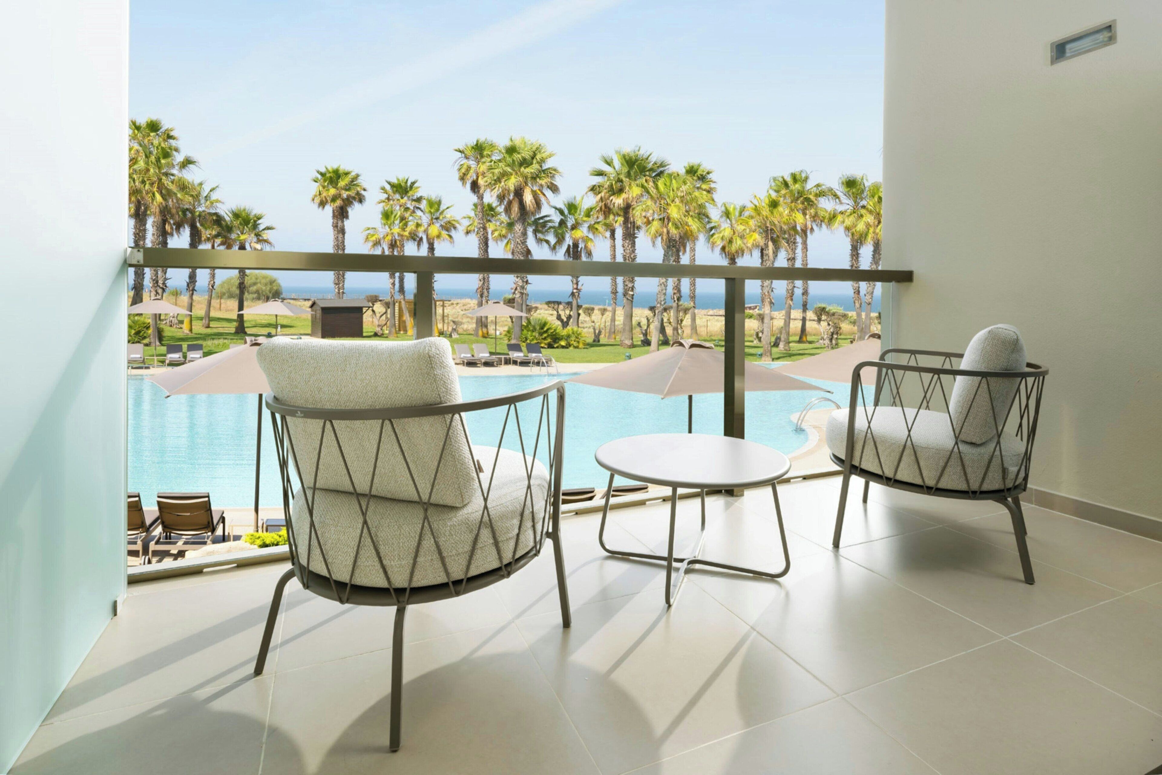 Un balcón con dos sillas y una mesa pequeña, con vista a una piscina y palmeras.
