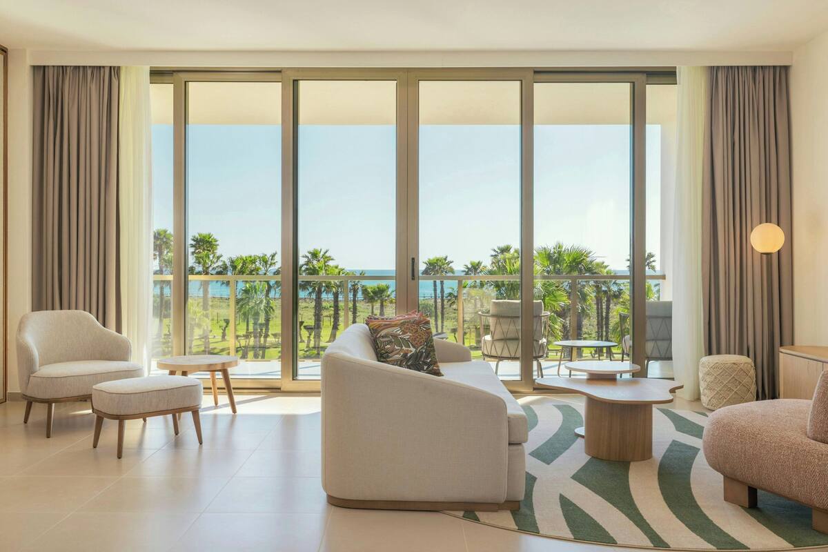 A spacious living room with a large window overlooking a beach, furnished with a sofa, chairs, and a coffee table.