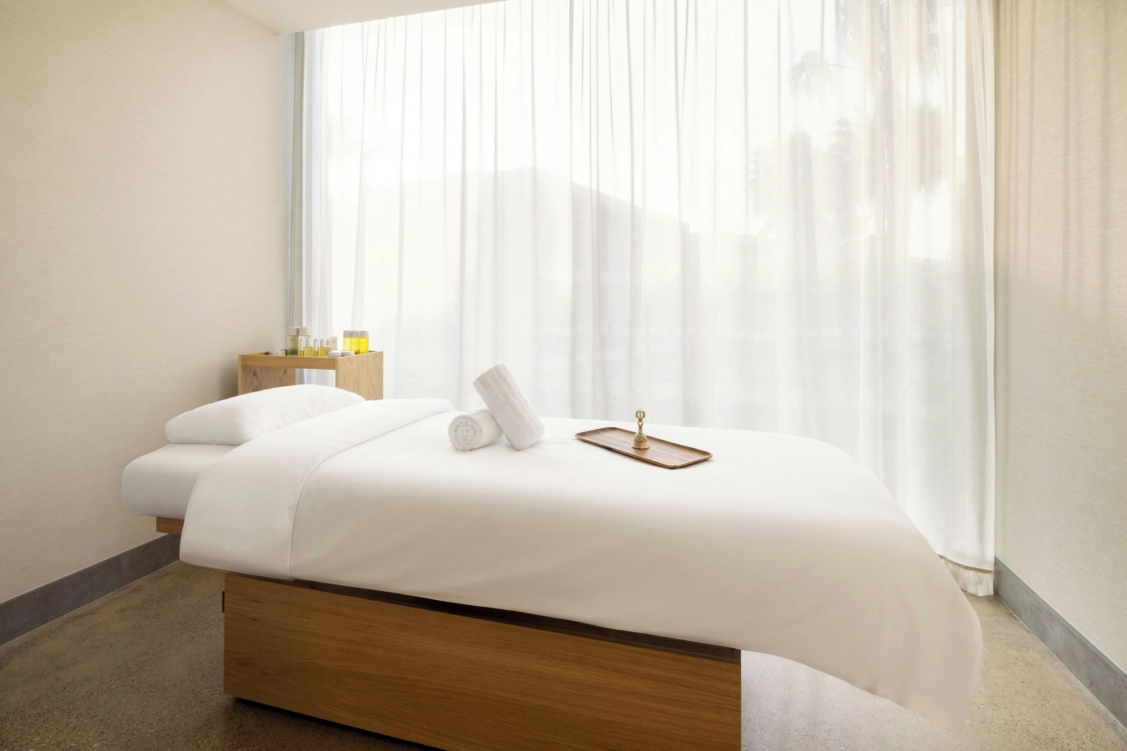 A modern hotel room with a wooden bed, white linens, a small bedside table with toiletries, and sheer curtains.