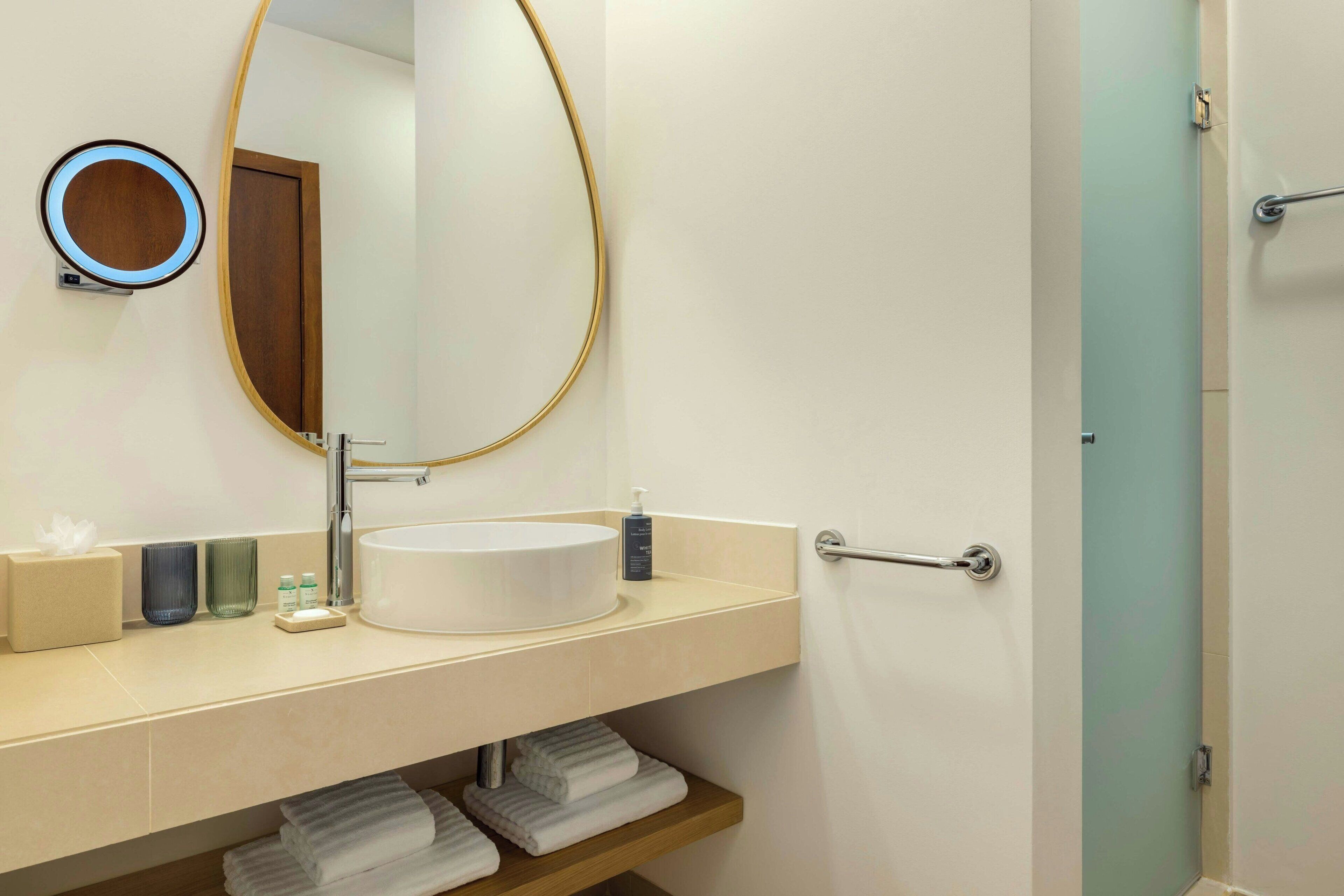 Un baño con un espejo redondo, un lavamanos, un perchero para toallas y una puerta de ducha.